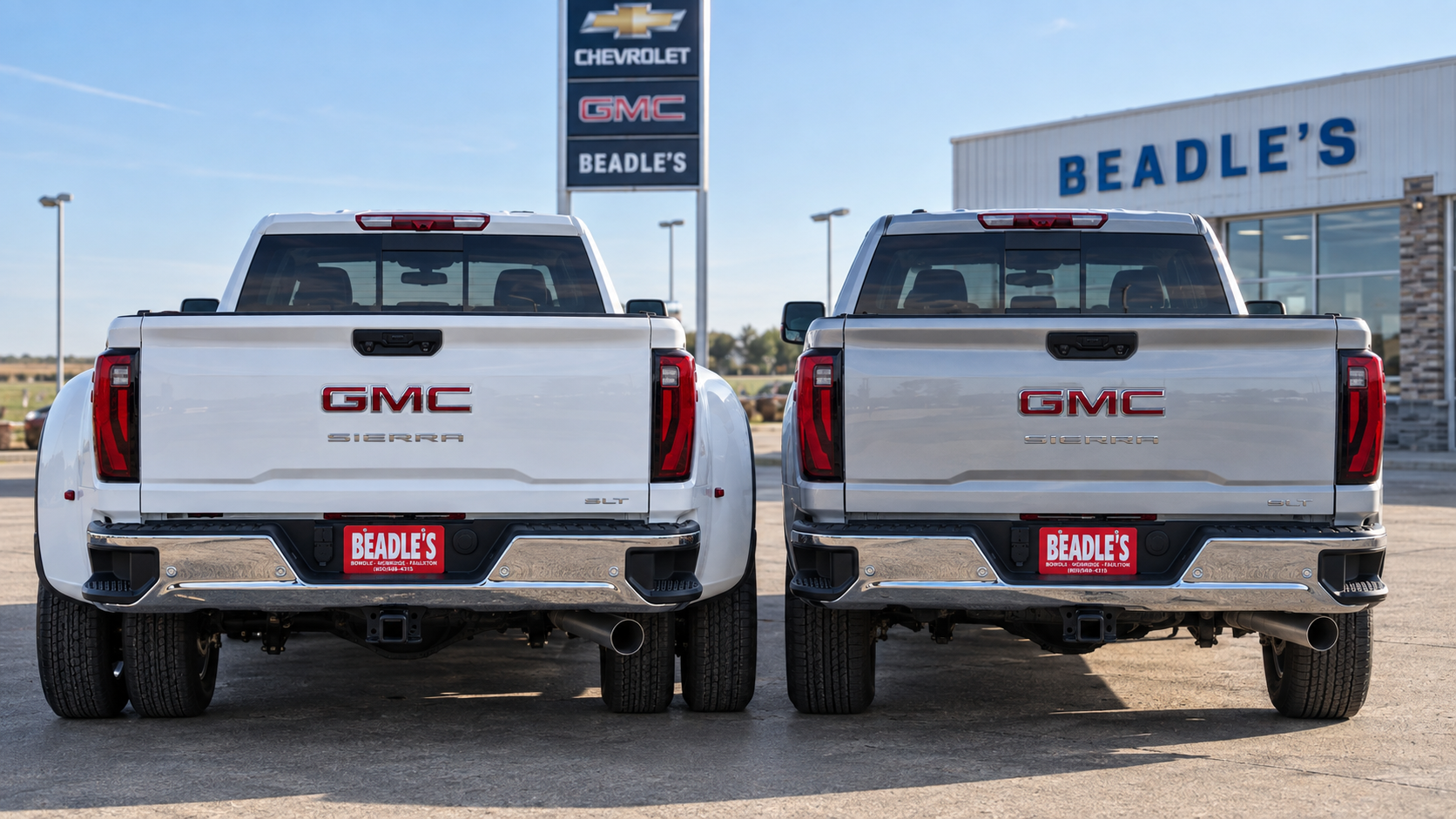 A 2026 GMC Sierra 3500 HD dually and a single-rear-wheel Sierra 3500 HD parked side by side, seen from the rear, at Beadle Chevrolet GMC in Mobridge, South Dakota