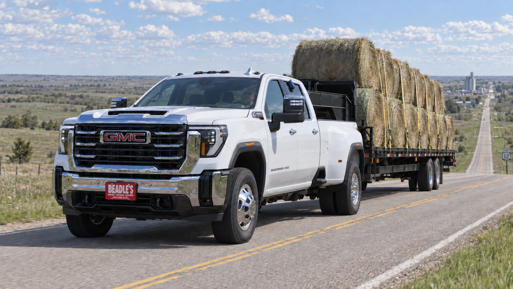 Summit White 2027 GMC Sierra 3500 HD dually pulling a gooseneck flatbed loaded with round bales up a US Highway 12 grade near Mobridge, South Dakota